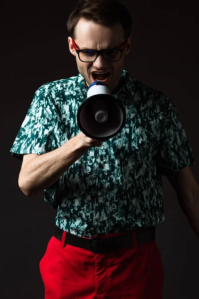 Fashionable man in eyeglasses in blue colorful shirt and red pants shouting in loudspeaker isolated on black — Stock Photo