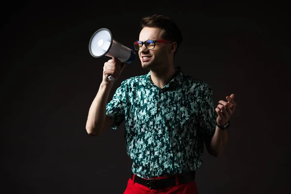 Homem na moda em óculos em camisa colorida azul e calças vermelhas gritando em alto-falante isolado em preto — Fotografia de Stock