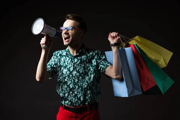 Uomo alla moda in occhiali in camicia colorata blu e pantaloni rossi con shopping bag urlando in altoparlante isolato su nero — Foto stock