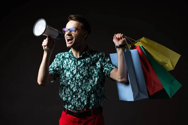 Homme à la mode dans les lunettes en chemise bleue colorée et pantalon rouge avec des sacs à provisions criant dans haut-parleur isolé sur noir — Photo de stock