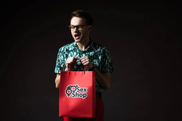 Fashionable man in eyeglasses in blue colorful shirt and red pants holding red shopping bag from sex shop isolated on black — Stock Photo