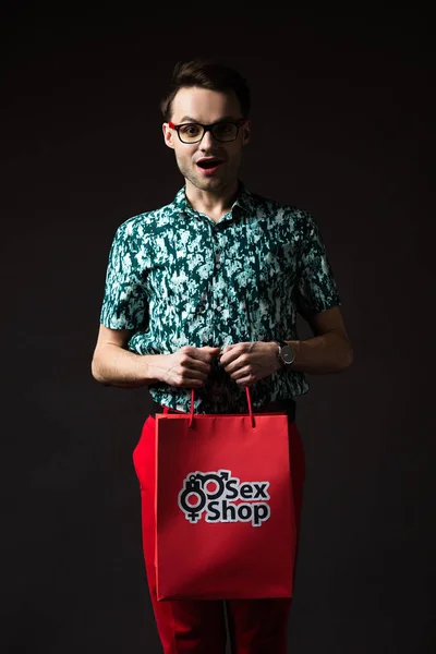Scioccato uomo alla moda in occhiali da vista in camicia colorata blu e pantaloni rossi in possesso di shopping bag rosso dal sexy shop isolato su nero — Foto stock