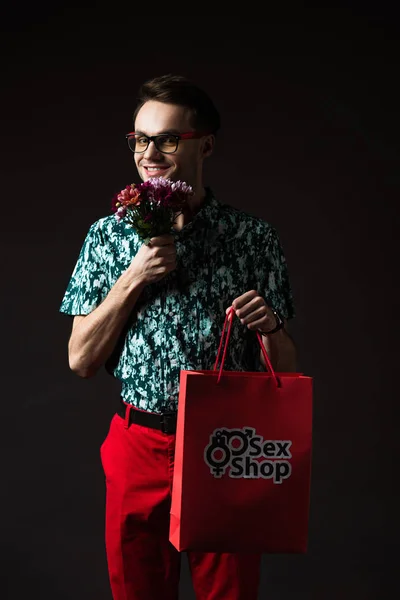 Uomo alla moda sorridente in occhiali da vista in camicia colorata blu e pantaloni rossi in possesso di shopping bag rosso dal sexy shop e fiori isolati su nero — Foto stock