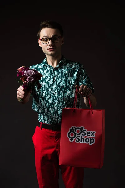 Uomo alla moda sorridente in occhiali da vista in camicia colorata blu e pantaloni rossi in possesso di shopping bag rosso dal sexy shop e fiori isolati su nero — Foto stock