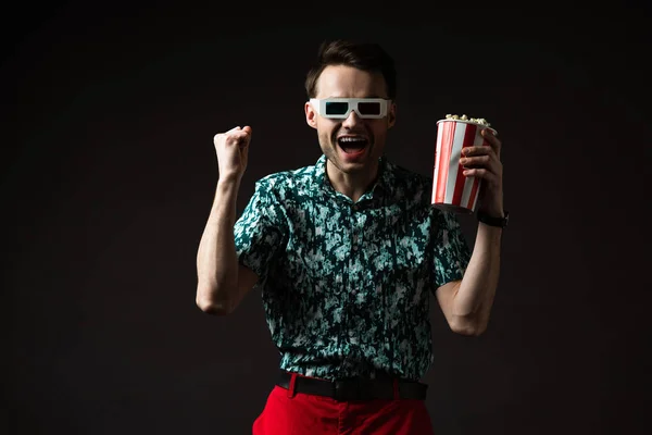 Excited fashionable man in 3d glasses in blue colorful shirt and red pants holding popcorn and showing yeah gesture isolated on black — Stock Photo