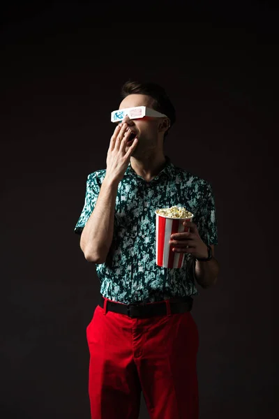 Hombre de moda en gafas 3d con boca abierta en camisa de colores azules y pantalones rojos sosteniendo palomitas de maíz aisladas en negro - foto de stock