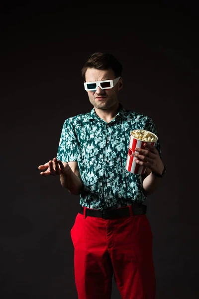 Confused fashionable man in 3d glasses in blue colorful shirt and red pants holding popcorn isolated on black — Stock Photo