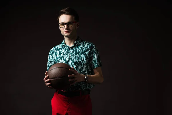 Homem na moda em óculos em azul camisa colorida e calças vermelhas segurando basquete isolado em preto — Fotografia de Stock