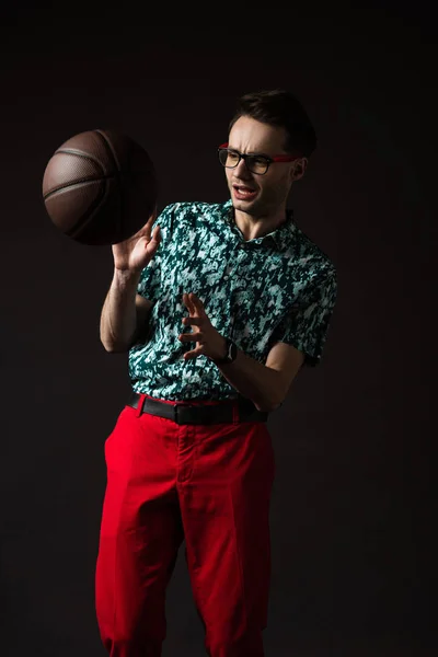 Homem elegante assustado em óculos em camisa colorida azul e calças vermelhas com basquete isolado em preto — Fotografia de Stock