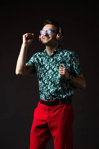 Homem na moda feliz em óculos ouvindo música em fones de ouvido e dançando isolado em preto — Fotografia de Stock