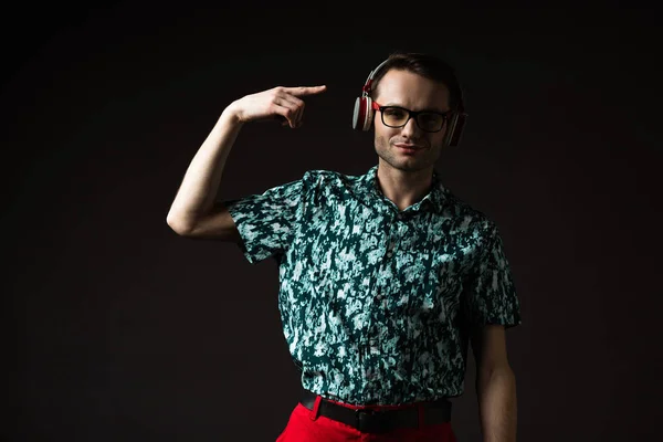 Hombre de moda en gafas que apuntan con el dedo a los auriculares aislados en negro - foto de stock