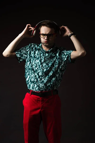 Homem na moda confuso em óculos ouvindo música em fones de ouvido isolados em preto — Fotografia de Stock