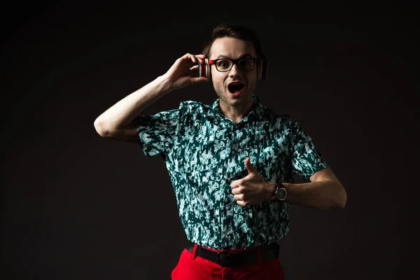 Shocked fashionable man in eyeglasses listening music in headphones and showing thumb up isolated on black — Stock Photo