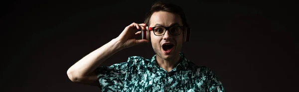 Aufgeregter modischer Mann mit Brille, der Musik in Kopfhörern hört, isoliert auf schwarzer, panoramischer Aufnahme — Stockfoto