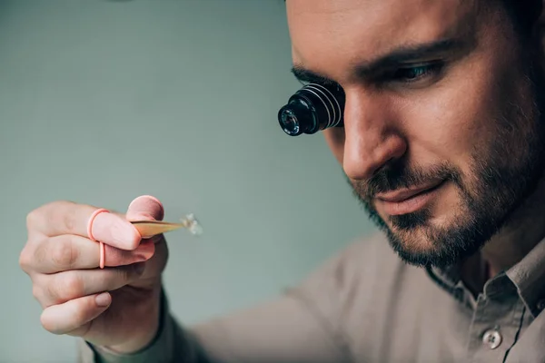 Handsome watchmaker in eyeglass loupe holding watch part isolated on grey — Stock Photo