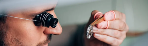 Panoramic shot of watchmaker in eyeglass loupe holding watch detail in tweezers — Stock Photo