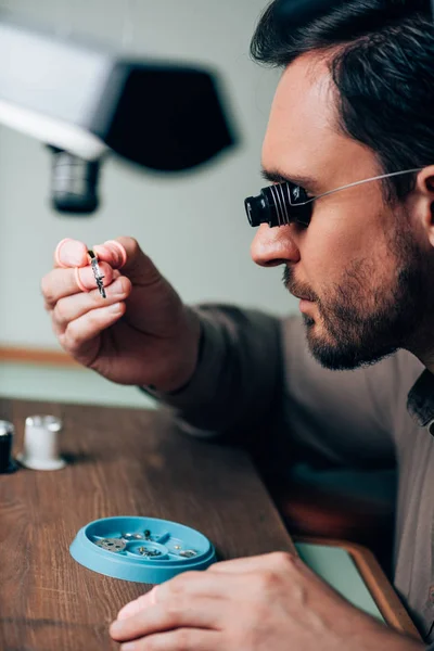 Side view of clockmaker in eyeglass tool holding watchpart in tweezers at table — Stock Photo