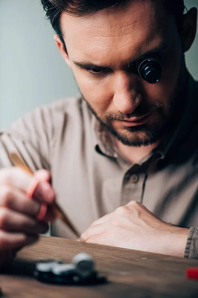 Selective focus of handsome watchmaker in eyeglass loupe working with tool tray and tweezers at table — Stock Photo