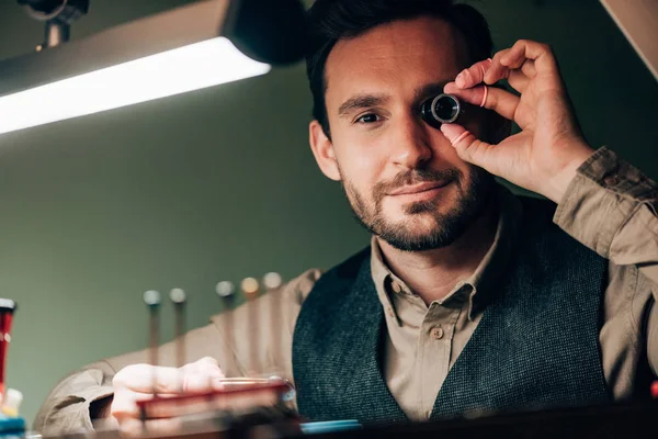 Selective focus of watchmaker holding eyeglass loupe and looking at camera by working table — Stock Photo