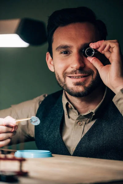 Sonriente relojero con lupa de anteojos sosteniendo parte del reloj mientras sostiene en la cámara - foto de stock