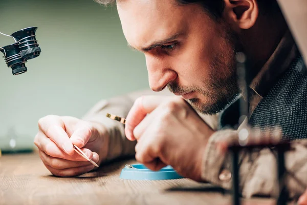 Side view of handsome watchmaker using magnifying glass for clock part at working table — Stock Photo