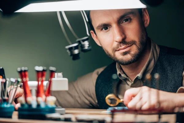 Horloger beau regardant la caméra tout en travaillant avec des pièces de montre et loupe — Photo de stock