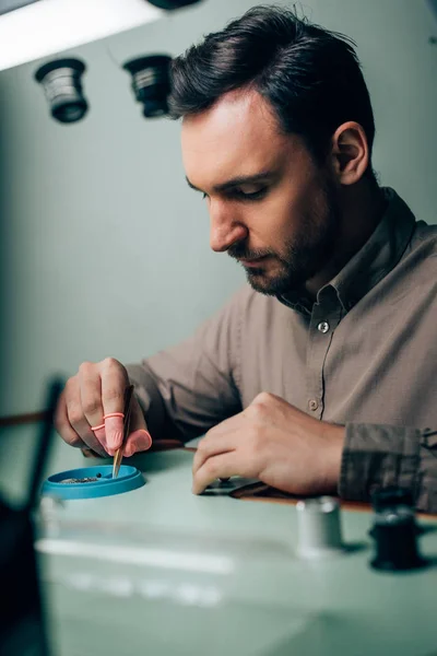 Vue latérale du bel horloger travaillant avec des pièces de montre par loupes de lunettes sur la table — Photo de stock