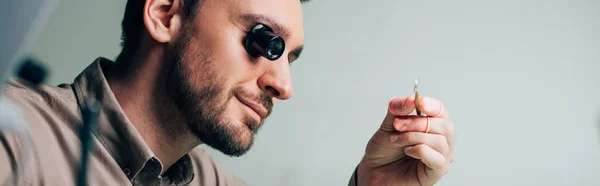 Panoramic shot of watchmaker in eyeglass loupe holding watch part — Stock Photo