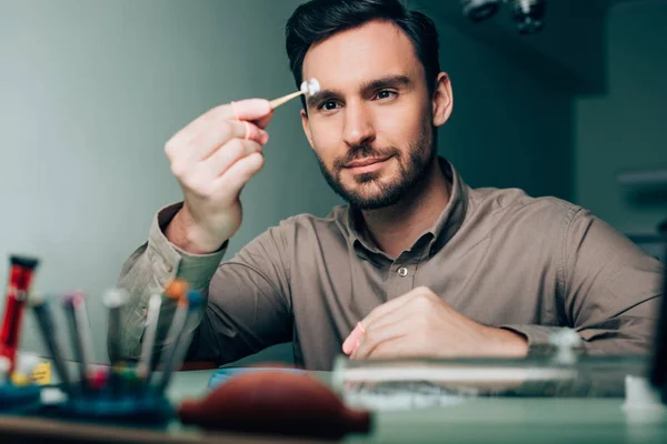 Selective focus of handsome watchmaker looking at watch part at working table — Stock Photo