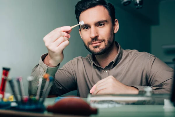 Enfoque selectivo del guapo relojero mirando la pieza del reloj en la mesa de trabajo - foto de stock