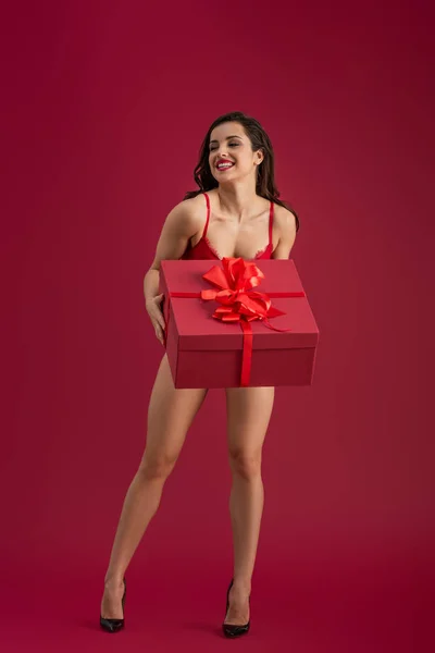 Sexy chica en lencería y zapatos de tacón alto sosteniendo gran caja de regalo mientras mira hacia otro lado y sonriendo sobre fondo rojo - foto de stock