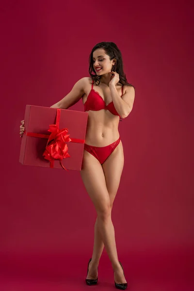 Sexy, chica sonriente en lencería y zapatos de tacón alto sosteniendo gran caja de regalo con cinta y lazo sobre fondo rojo - foto de stock