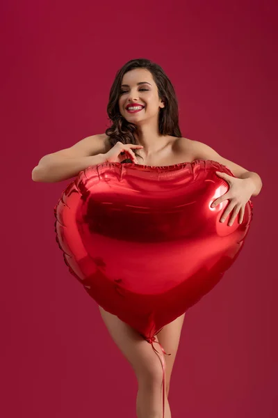 Chica seductora sonriendo y mirando hacia otro lado mientras sostiene un gran globo en forma de corazón aislado en rojo - foto de stock