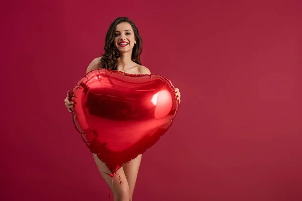 Chica sexy sonriendo a la cámara mientras sostiene el globo en forma de corazón aislado en rojo - foto de stock