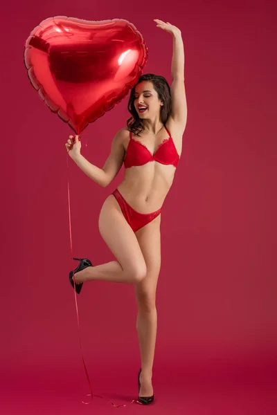 Sexy, menina alegre em lingerie e sapatos de salto alto segurando balão em forma de coração, enquanto em pé em uma perna no fundo vermelho — Fotografia de Stock