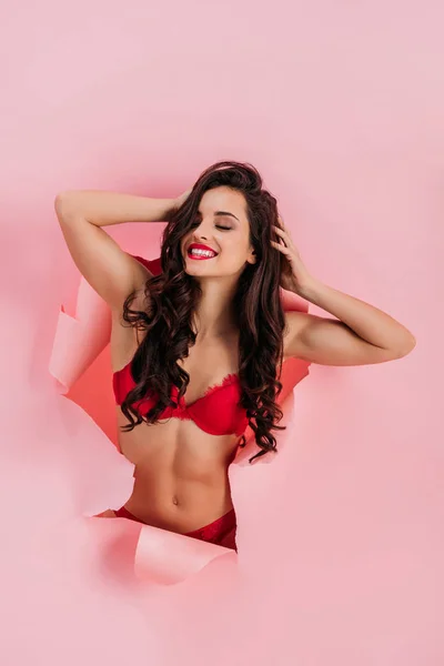Сексуальна, весела дівчина в червоній білизні, що розтягується закритими очима в паперовій дірі на рожевому фоні — стокове фото