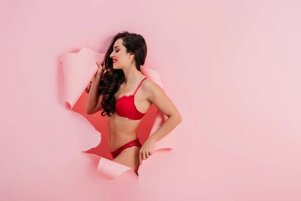 Séduisante, fille souriante en lingerie rouge debout dans un trou de papier sur fond rose — Photo de stock