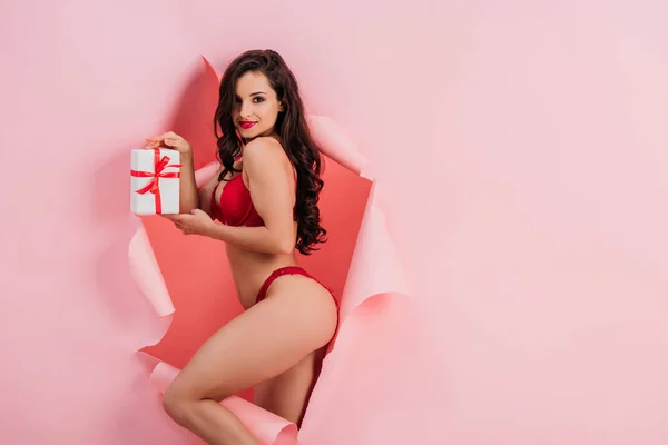 Passionate girl in red lingerie holding gift box and smiling at camera while standing in paper hole on pink background — Stock Photo