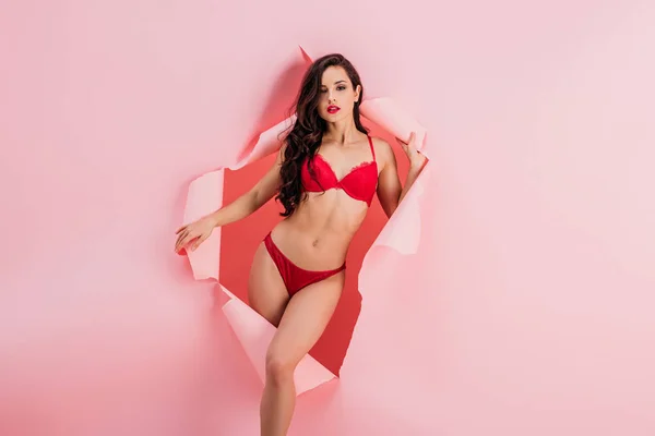 Seductive girl in red lingerie looking at camera while posing in paper hole on pink background — Stock Photo