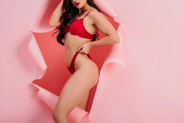 Vista recortada de chica sexy en ropa interior roja tocando bragas mientras está de pie en el agujero de papel sobre fondo rosa - foto de stock