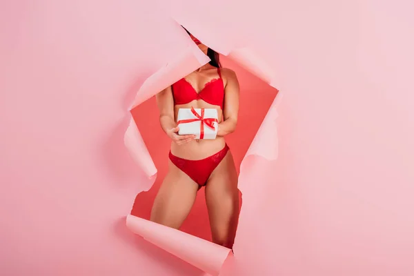 Vue recadrée de fille sexy en lingerie tenant boîte cadeau tout en se tenant dans un trou de papier sur fond rose — Photo de stock