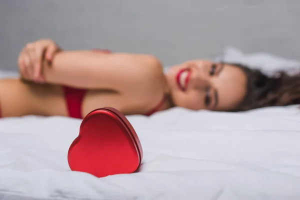 Selective focus of heart-shaped gift box near sexy, smiling girl lying in bed — Stock Photo