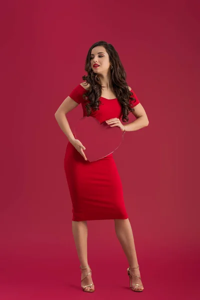 Menina sexy em vestido vermelho elegante e sandálias de salto alto segurando coração de papel enquanto em pé isolado no vermelho — Fotografia de Stock