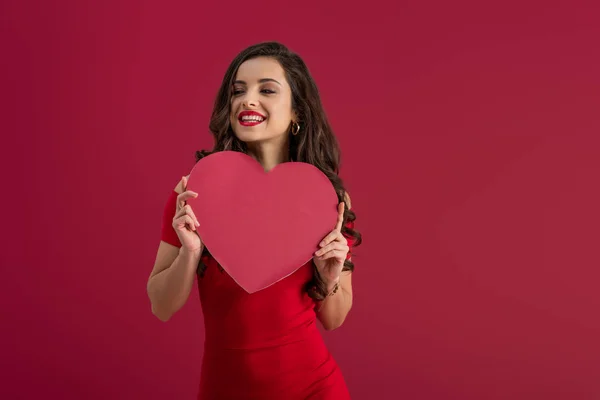 Sensual, elegante chica sosteniendo el corazón de papel mientras sonríe aislado en rojo - foto de stock