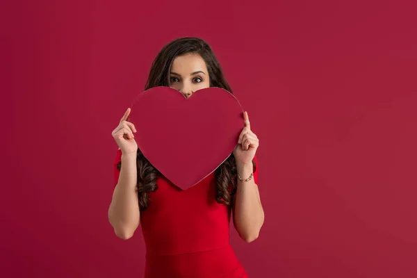 Sexy, elegant girl covering face with paper heart while looking at camera isolated on red — Stock Photo