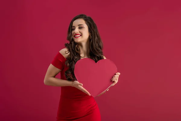 Sensual, elegante chica sosteniendo el corazón de papel mientras sonríe aislado en rojo - foto de stock