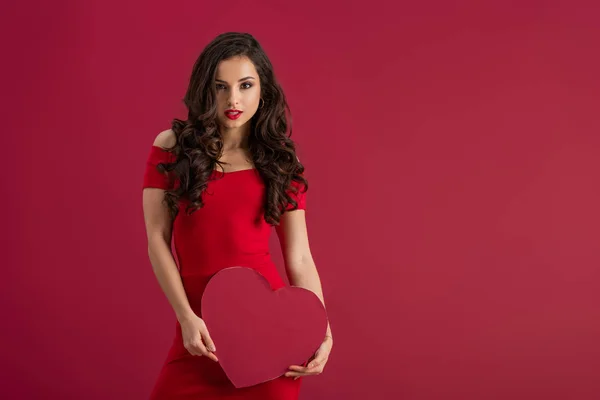 Seductive, elegant girl holding paper heart while looking at camera isolated on red — Stock Photo