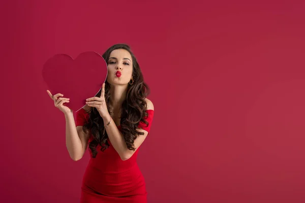 Сексуальная, элегантная девушка с бумажным сердцем, отправляя воздушный поцелуй в камеру, изолированную на красный — стоковое фото