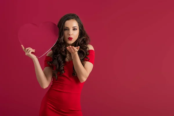 Kokette, elegante Mädchen mit Papierherz, während sie Luftküsse in die Kamera sendet, isoliert auf Rot — Stockfoto