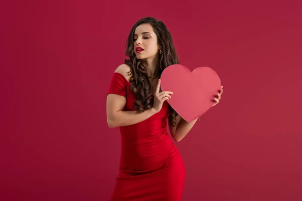 Seductora y elegante niña sosteniendo el corazón de papel mientras está de pie con los ojos cerrados aislados en rojo - foto de stock